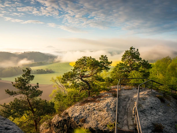Uitzicht vanaf de Keizerskroon Rotsachtige uitloper met hekwerk, omringd door groene bomen, met uitzicht op mistige velden en heuvels in het ochtendlicht.
