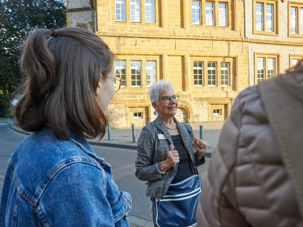 Bielefeld-Ratsgymnasium-Stadtführung-Teutoburger-Wald-Tourismus-T-Evers-094.jpg Eine ältere Frau führt eine Gruppe bei einer Stadtführung vor einem historischen Gebäude in Bielefeld.