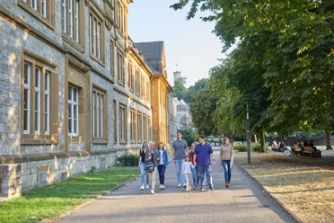Bielefeld-Ratsgymnasium-Stadtführung-Teutoburger-Wald-Tourismus-T-Evers-104.jpg Sechs Personen spazieren auf einem Weg neben einem historischen Gebäude, umgeben von Bäumen.