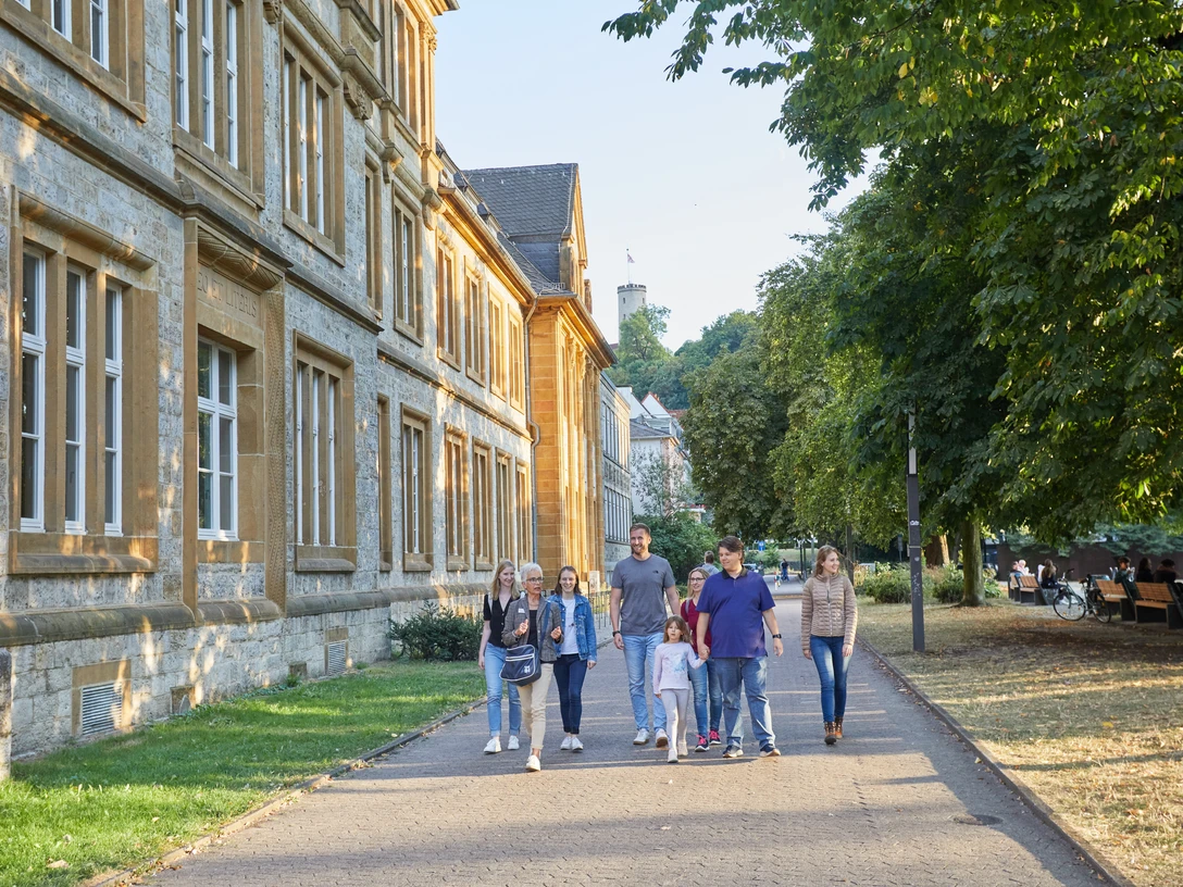 Bielefeld-Ratsgymnasium-Stadtführung-Teutoburger-Wald-Tourismus-T-Evers-104.jpg Sechs Personen spazieren auf einem Weg neben einem historischen Gebäude, umgeben von Bäumen.