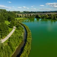 Flusslandschaft mit Brücke im Hintergrund, Radfahrer auf Weg; grüne Natur unter blauem Himmel.