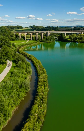 Flusslandschaft mit Brücke im Hintergrund, Radfahrer auf Weg; grüne Natur unter blauem Himmel.