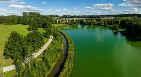 Bielefeld-Obersee-Teutoburger-Wald-Tourismus-D-Ketz-063.jpg Flusslandschaft mit Brücke im Hintergrund, Radfahrer auf Weg; grüne Natur unter blauem Himmel.