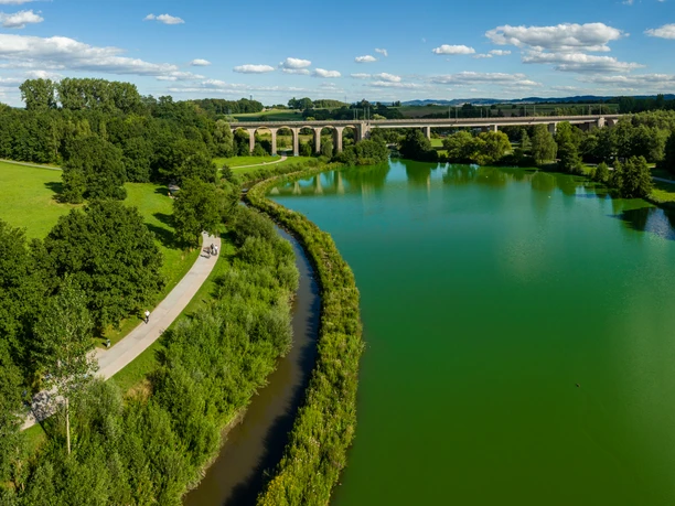Bielefeld-Obersee-Teutoburger-Wald-Tourismus-D-Ketz-063.jpg Flusslandschaft mit Brücke im Hintergrund, Radfahrer auf Weg; grüne Natur unter blauem Himmel.