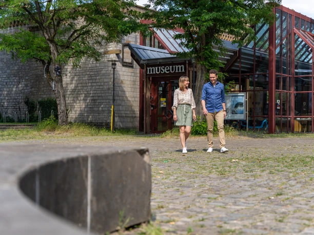 Bielefeld-RavensbergerPark-Teutoburger-Wald-Tourismus-Patrick-Gawandtka-080.jpg <p>Ein Paar spaziert draußen vor einem modernen Glasgebäude mit Museum-Logo, umgeben von Bäumen.</p>