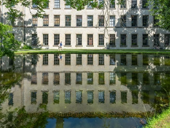 Alter Klinikbau spiegelt sich im ruhigen Teich, drei Menschen spazieren am sonnigen Ufer entlang.