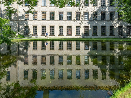 Alter Klinikbau spiegelt sich im ruhigen Teich, drei Menschen spazieren am sonnigen Ufer entlang.