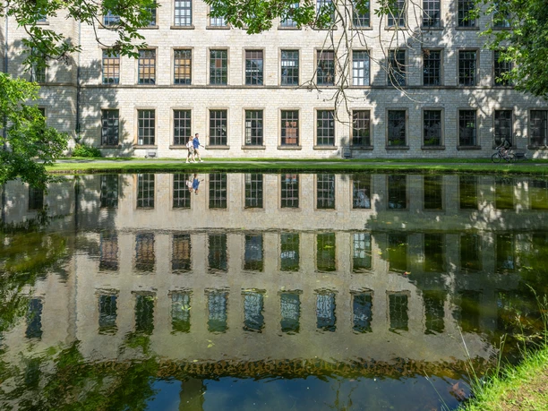 Alter Klinikbau spiegelt sich im ruhigen Teich, drei Menschen spazieren am sonnigen Ufer entlang.