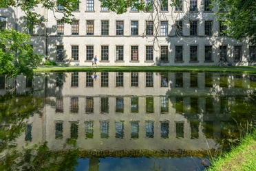 Bielefeld-RavensbergerPark-Teutoburger-Wald-Tourismus-Patrick-Gawandtka-084.jpg Alter Klinikbau spiegelt sich im ruhigen Teich, drei Menschen spazieren am sonnigen Ufer entlang.