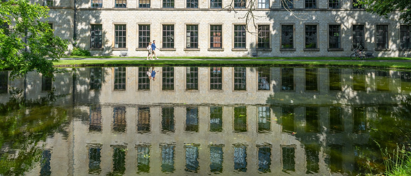 Bielefeld-RavensbergerPark-Teutoburger-Wald-Tourismus-Patrick-Gawandtka-084.jpg Alter Klinikbau spiegelt sich im ruhigen Teich, drei Menschen spazieren am sonnigen Ufer entlang.