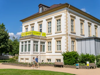 Bielefeld-RavensbergerPark-Teutoburger-Wald-Tourismus-Patrick-Gawandtka-072.jpg <p>Hellgelbes, zweistöckiges Museum, flankiert von Bäumen, mit Spaziergängern im Vordergrund.</p>