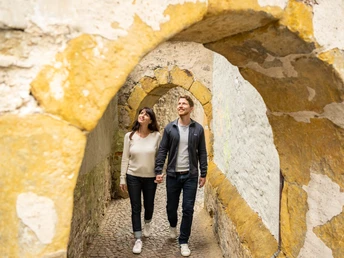 Zwei Personen spazieren lächelnd durch einen historischen, steinernen Torbogen.