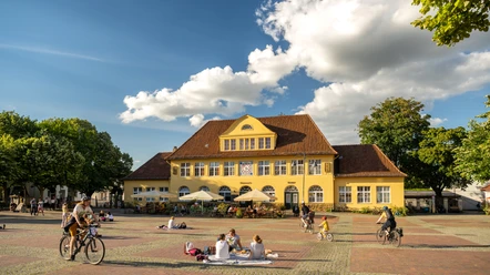 Bielefeld-Siegfriedplatz (Siggi)-Teutoburger-Wald-Tourismus-D-Ketz-087.jpg Gelbes Gebäude im ländlichen Stil vor blauem Himmel, Menschen genießen sonnigen Platz, einige radfahren.