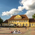 Bielefeld-Siegfriedplatz (Siggi)-Teutoburger-Wald-Tourismus-D-Ketz-087.jpg Gelbes Gebäude im ländlichen Stil vor blauem Himmel, Menschen genießen sonnigen Platz, einige radfahren.