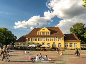 Bielefeld-Siegfriedplatz (Siggi)-Teutoburger-Wald-Tourismus-D-Ketz-087.jpg Gelbes Gebäude im ländlichen Stil vor blauem Himmel, Menschen genießen sonnigen Platz, einige radfahren.
