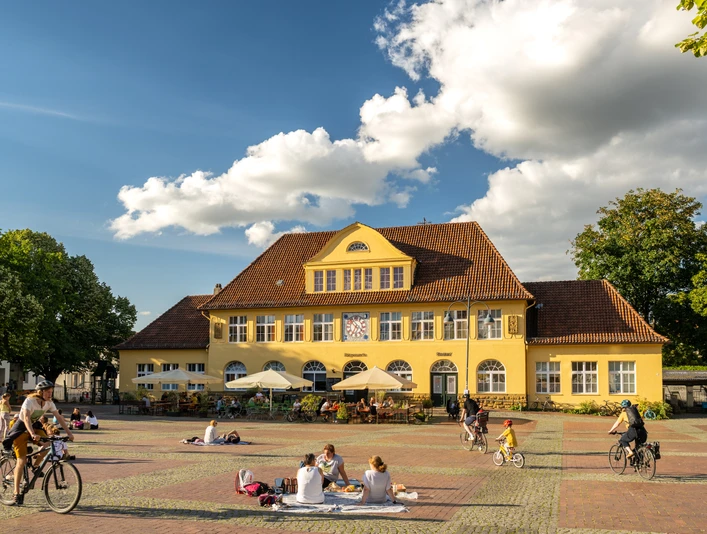 Bielefeld-Siegfriedplatz (Siggi)-Teutoburger-Wald-Tourismus-D-Ketz-087.jpg Gelbes Gebäude im ländlichen Stil vor blauem Himmel, Menschen genießen sonnigen Platz, einige radfahren.