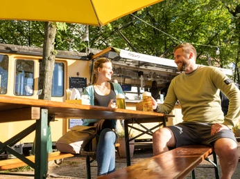 Bielefeld-Siegfriedplatz (Siggi)-Teutoburger-Wald-Tourismus-D-Ketz-090.jpg Pärchen genießt Bier unter Sonnenschirm vor gelbem Imbisswagen in grüner, sonniger Umgebung.