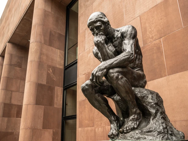 Das Bild zeigt die Statue von Auguste Rodins "Der Denker" vor einem roten Sandsteingebäude.