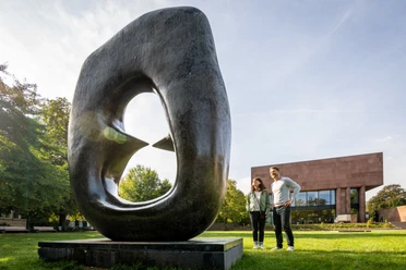 Skulptur mit Loch, im Vordergrund lächelndes Paar, moderne Architektur im Hintergrund, sonniger Tag.