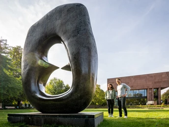 Skulptur mit Loch, im Vordergrund lächelndes Paar, moderne Architektur im Hintergrund, sonniger Tag.