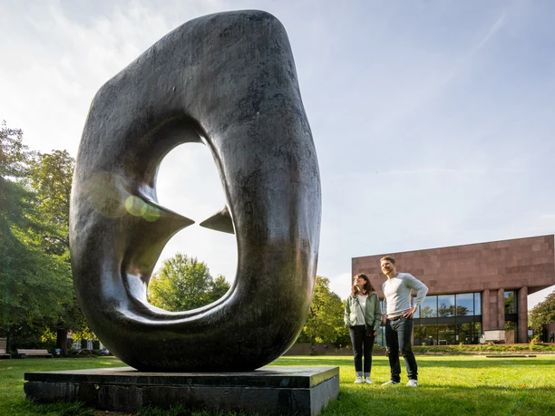 Bielefeld-Skulpturenpark-Teutoburger-Wald-Tourismus-D-Ketz-055.jpg Skulptur mit Loch, im Vordergrund lächelndes Paar, moderne Architektur im Hintergrund, sonniger Tag.