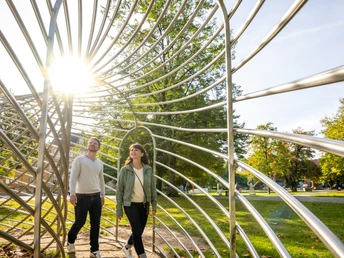 <p>Ein Paar spaziert durch eine skulpturale Metallstruktur im Park, bei Sonnenschein und Bäumen im Hintergrund.</p>