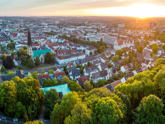 Bielefeld-Sparrenburg-Teutoburger-Wald-Tourismus-D-Ketz-012.jpg <p>Luftaufnahme einer Stadt bei Sonnenuntergang mit Kirche, Wohnhäusern und Bäumen im Vordergrund.</p>