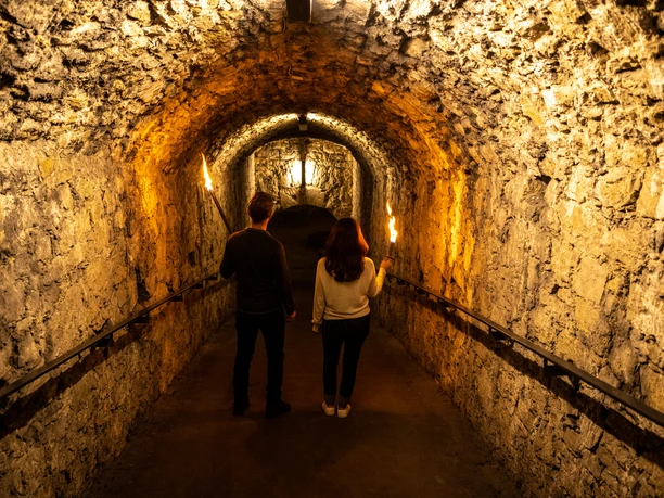 Bielefeld-Sparrenburg-Teutoburger-Wald-Tourismus-D-Ketz-024-CC-BY-SA.jpg Zwei Personen erkunden einen beleuchteten, steinernen Tunnel mit Fackeln in den Händen.