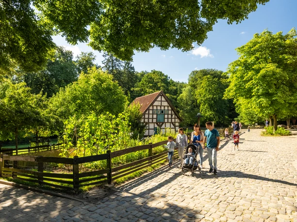 Historisches Fachwerkhaus im Grünen, umgeben von Spaziergängern auf einem sonnigen Kopfsteinpflasterweg.