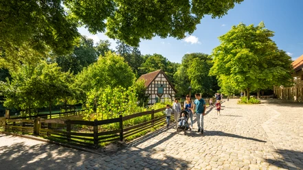 Bielefeld-Tierpark Olderdissen-Teutoburger-Wald-Tourismus-D-Ketz-024.jpg Historisches Fachwerkhaus im Grünen, umgeben von Spaziergängern auf einem sonnigen Kopfsteinpflasterweg.