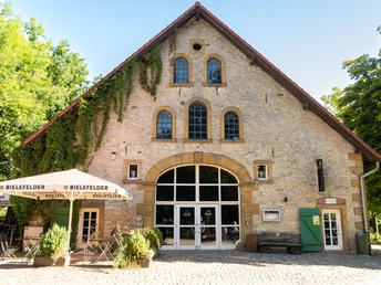 Bielefeld-Tierpark Olderdissen-Teutoburger-Wald-Tourismus-D-Ketz-004.jpg Altes Gebäude mit Steinfront, großen Fenstern, grünem Tor und Außenterrasse mit weißen Sonnenschirmen.
