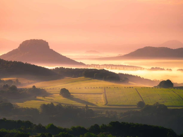 Blick vom Kleinen Bärenstein Sanfte Hügel und Felder im Morgennebel, beleuchtet von einem warmen, orangefarbenen Sonnenaufgang, mit einem markanten Berg im Hintergrund.
