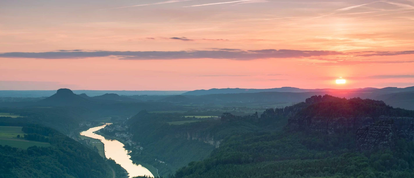 Pohled z Kipphorn View Západ slunce nad říční krajinou se zelenými kopci a jasnou oblohou, která září teplými pastelovými barvami.