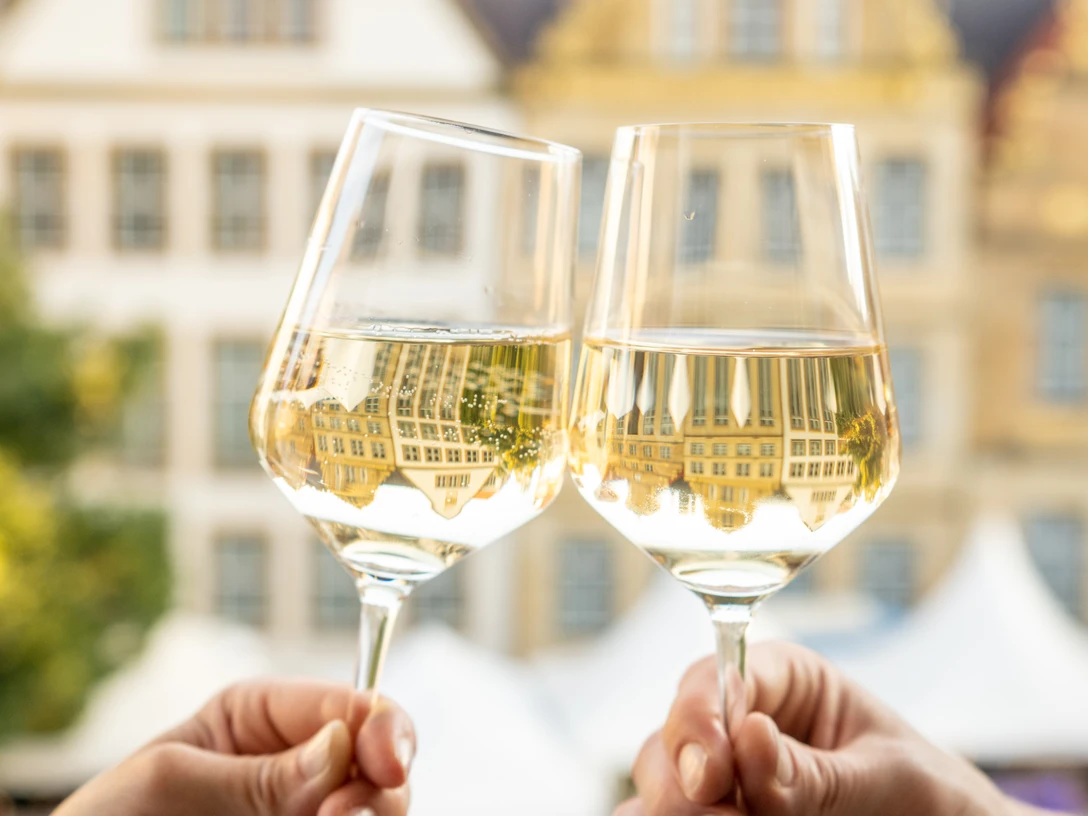 Bielefeld-Weinmarkt-Teutoburger-Wald-Tourismus-D-Ketz-093-CC-BY-SA.jpg Zwei Hände stoßen mit gefüllten Weingläsern vor historischem Gebäudehintergrund an.