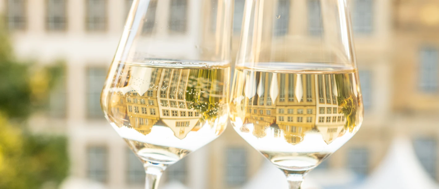 Bielefeld-Weinmarkt-Teutoburger-Wald-Tourismus-D-Ketz-093-CC-BY-SA.jpg Zwei Hände stoßen mit gefüllten Weingläsern vor historischem Gebäudehintergrund an.