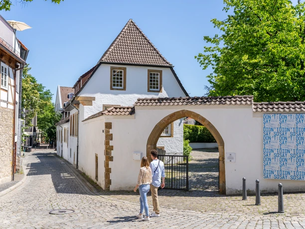 <p>Pärchen spaziert auf gepflasterter Gasse zu historischem Torbogen in sonniger Altstadt.</p>