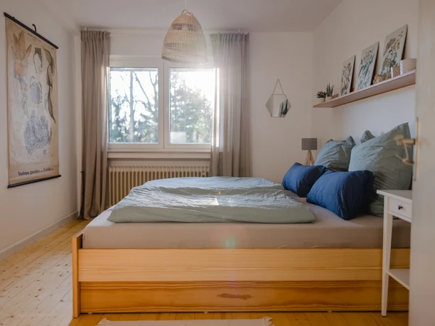 Waldferienwohnung_Schlafzimmer_Katharina_Hein.JPG Helles Schlafzimmer mit Holzboden, einem großen Doppelbett mit blauen Kissen und einem Fensterblick ins Grüne.