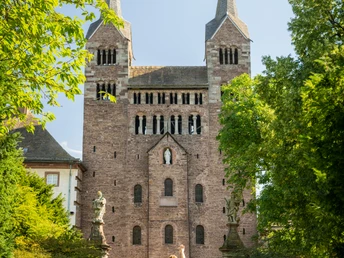 Hoexter-Corvey-Teutoburger-Wald-Tourismus-D-Ketz-040-CC-BY-SA.jpg Ein Paar spaziert auf einem gepflasterten Weg auf eine historische Backsteinkirche mit zwei Türmen zu.