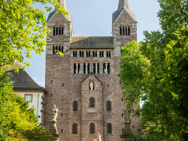 Hoexter-Corvey-Teutoburger-Wald-Tourismus-D-Ketz-040-CC-BY-SA.jpg Ein Paar spaziert auf einem gepflasterten Weg auf eine historische Backsteinkirche mit zwei Türmen zu.