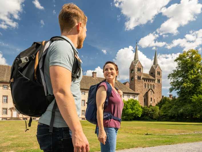 Hoexter-Corvey-Teutoburger-Wald-Tourismus-D-Ketz-074.jpg Ein Paar mit Rucksäcken spaziert lächelnd vor einer historischen Kathedrale mit zwei Türmen.