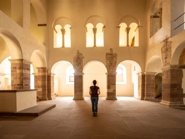 Eine Person steht in einer lichtdurchfluteten, historischen Halle mit Rundbögen und antiken Säulen.