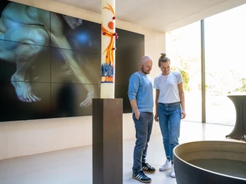 Mann und Frau betrachten Skulptur in modernem, lichtdurchflutetem Ausstellungsraum mit Bild im Hintergrund.
