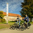 Zwei Radfahrer vor einem historischen Gebäude, umgeben von grünen Bäumen und blauen Himmel.