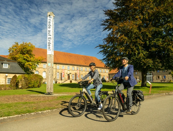 Zwei Radfahrer vor einem historischen Gebäude, umgeben von grünen Bäumen und blauen Himmel.