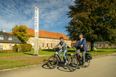 Warburg-Kloster HardehausenTeutoburger-Wald-Tourismus-D-Ketz-064.jpg Zwei Radfahrer vor einem historischen Gebäude, umgeben von grünen Bäumen und blauen Himmel.