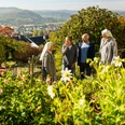 Blühender Garten mit vier Personen im Gespräch, darunter zwei Nonnen, umgeben von herbstlicher Landschaft.