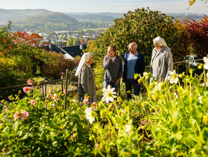 Warburg-Scherfede-Zionsberg-Teutoburger-Wald-Tourismus-D-Ketz-042.jpg Blühender Garten mit vier Personen im Gespräch, darunter zwei Nonnen, umgeben von herbstlicher Landschaft.