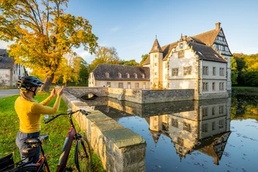 Willebadessen-Wasserschloss Schweckhausen-Teutoburger-Wald-Tourismus-D-Ketz-118.jpg Eine Radfahrerin fotografiert ein malerisches Wasserschloss umgeben von Bäumen und einem Graben.