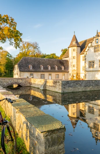 Willebadessen-Wasserschloss Schweckhausen-Teutoburger-Wald-Tourismus-D-Ketz-118.jpg Eine Radfahrerin fotografiert ein malerisches Wasserschloss umgeben von Bäumen und einem Graben.