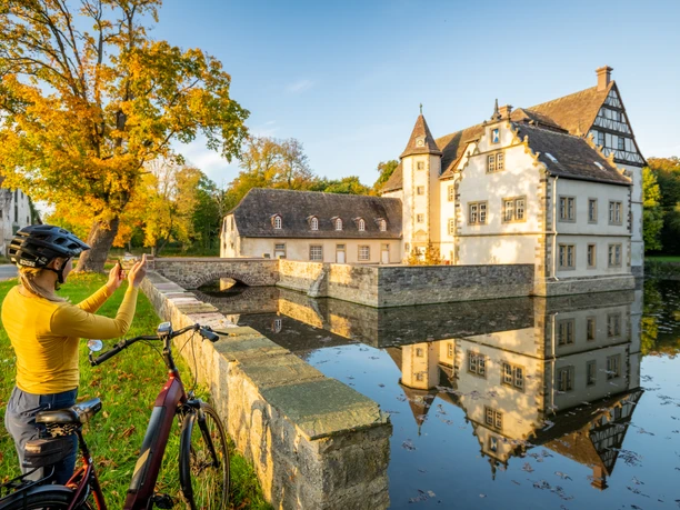 Willebadessen-Wasserschloss Schweckhausen-Teutoburger-Wald-Tourismus-D-Ketz-118.jpg Eine Radfahrerin fotografiert ein malerisches Wasserschloss umgeben von Bäumen und einem Graben.